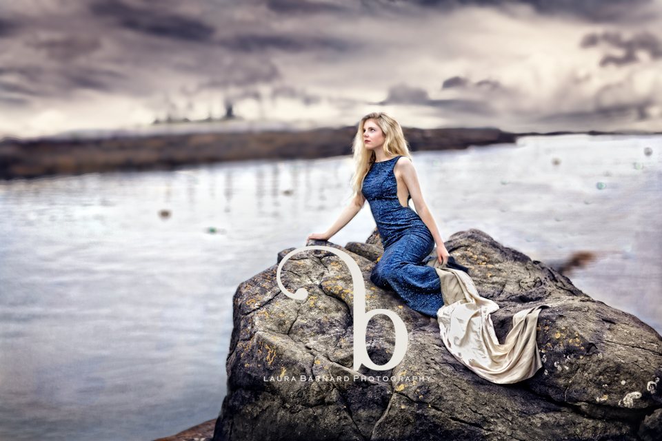 A senior portrait in a flowing blue gown on dark coastal rocks at dusk.