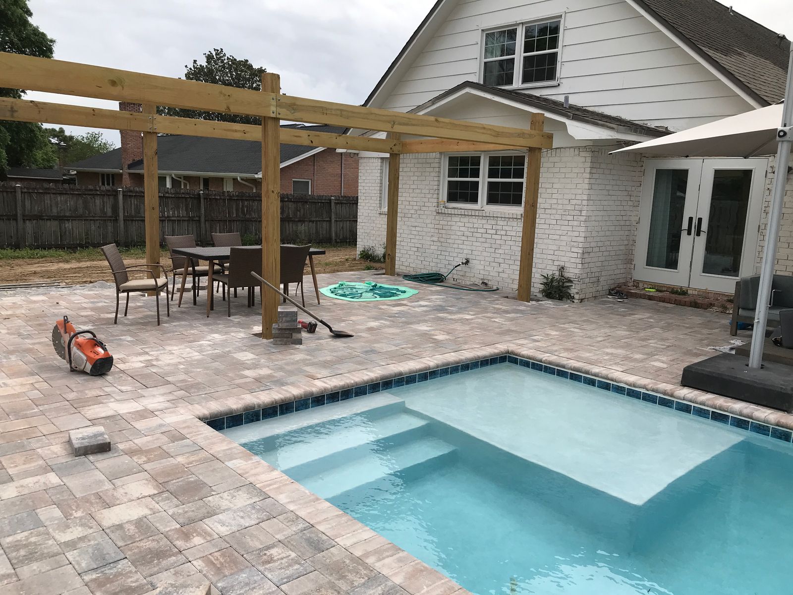 After: a finished paver pool deck with a wooden pergola, patio table and chairs